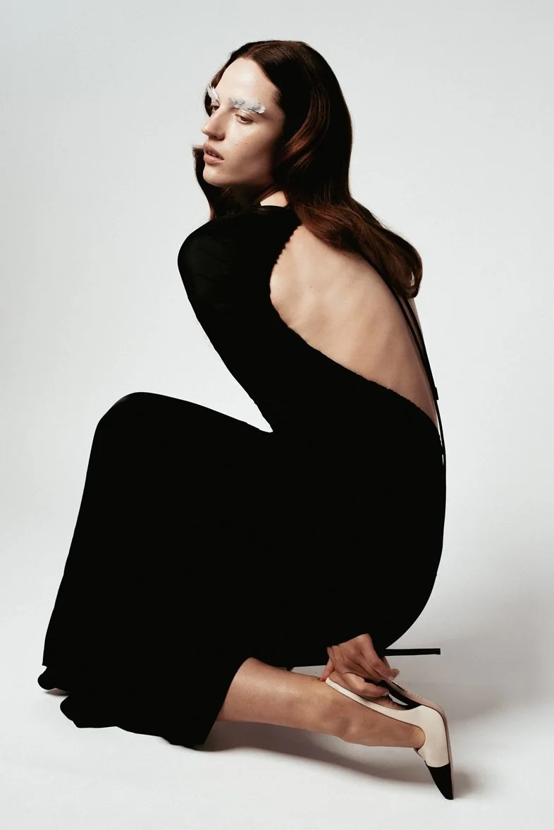 A woman with long, dark brown hair and white eyebrows is kneeling on the floor, wearing a black dress with a low, open back and white high heels. She is posing against a plain light-colored background.
