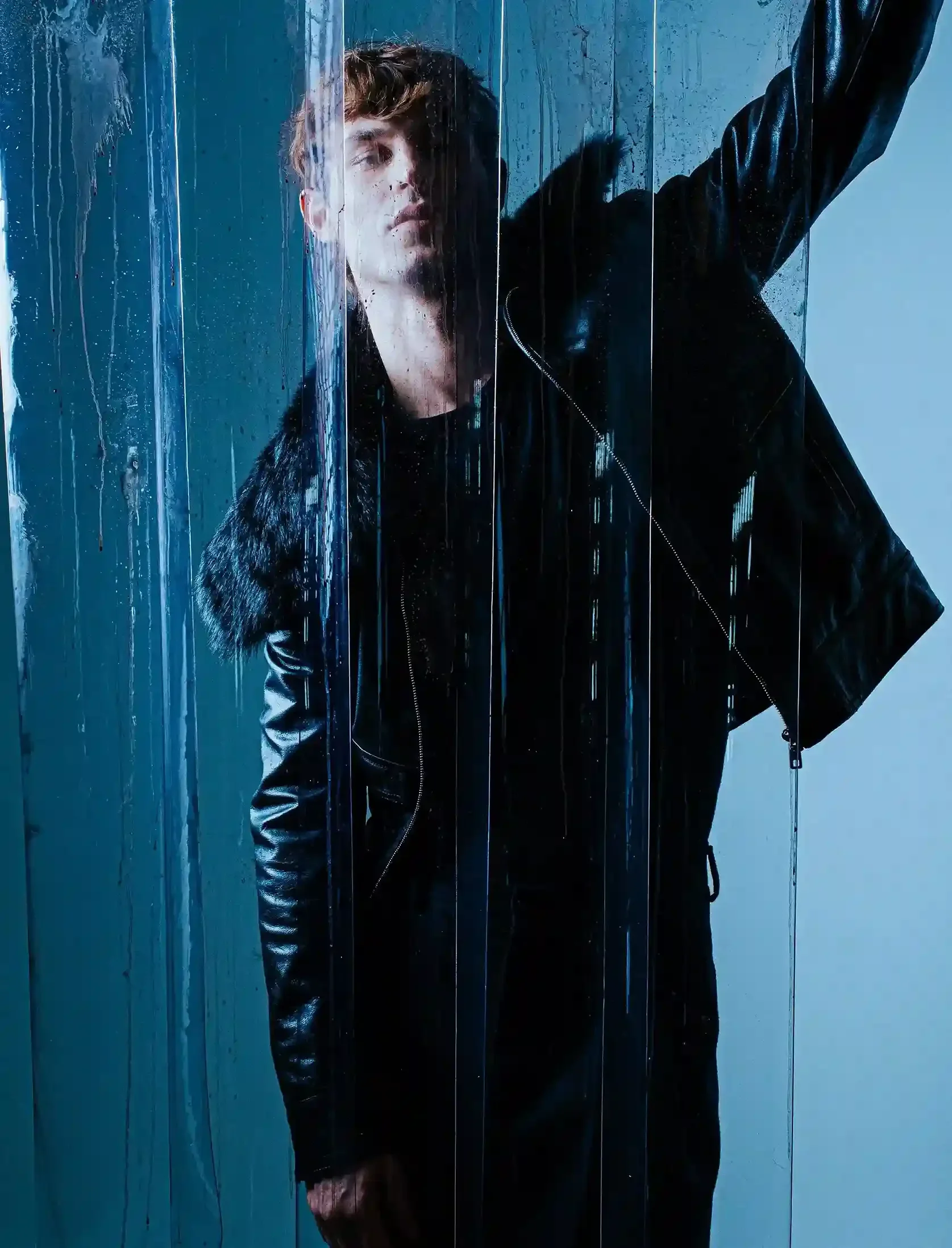 A young man with curly brown hair and a serious expression stands behind a rain-streaked glass wall, wearing a black leather jacket and a dark shirt, with his right arm raised.