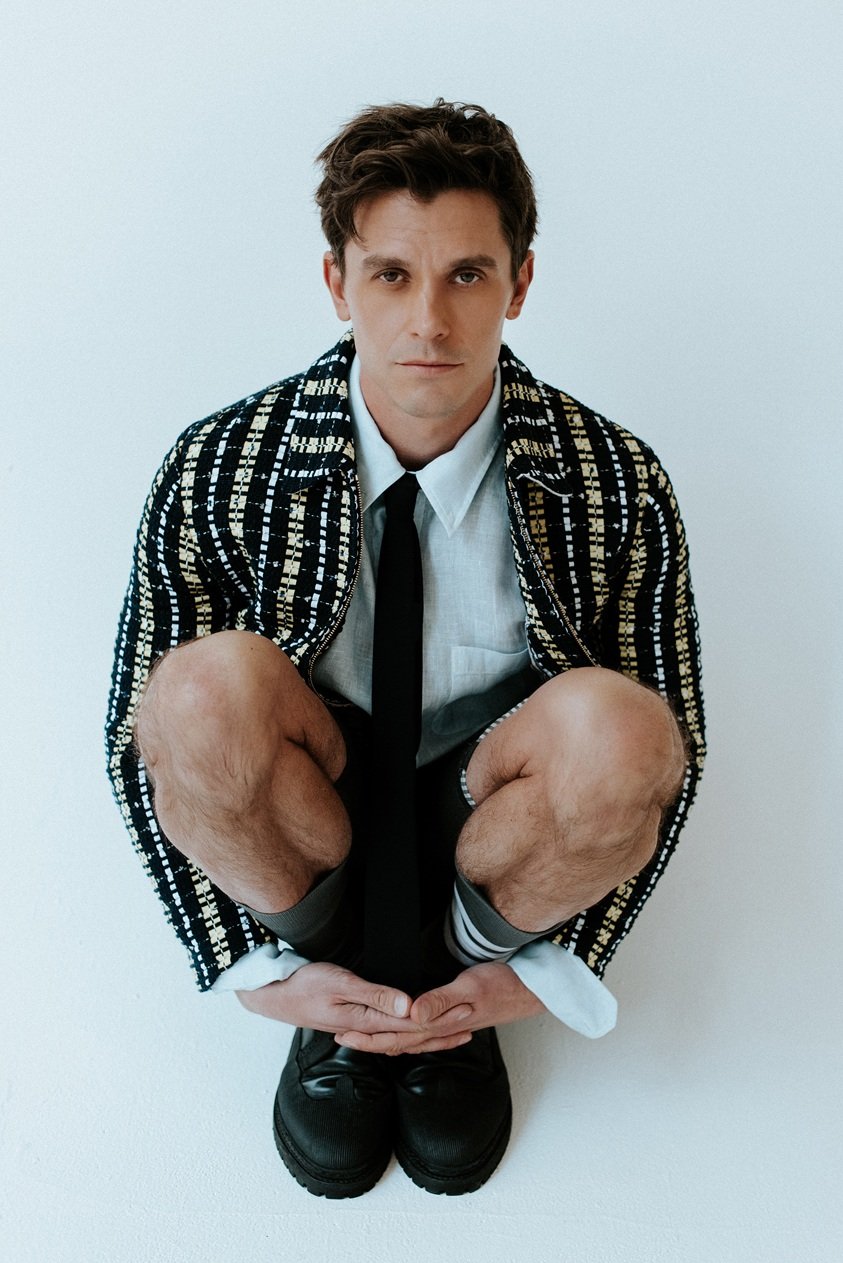 A man with dark hair, wearing a patterned jacket, white shirt, and black tie, crouches with knees bent and hands together, against a plain white background.