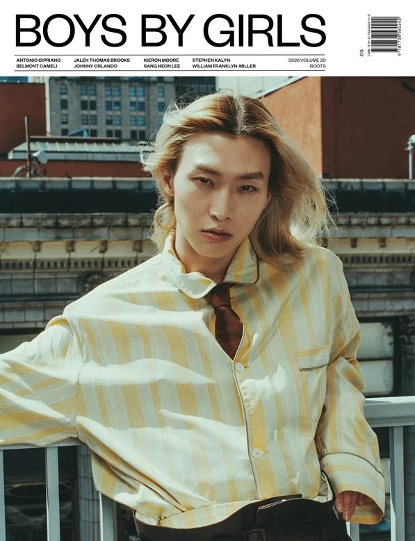 A person with long, wavy, blonde hair standing on a rooftop in an urban area, wearing a yellow and white striped shirt with a brown tie, against a backdrop of city buildings.
