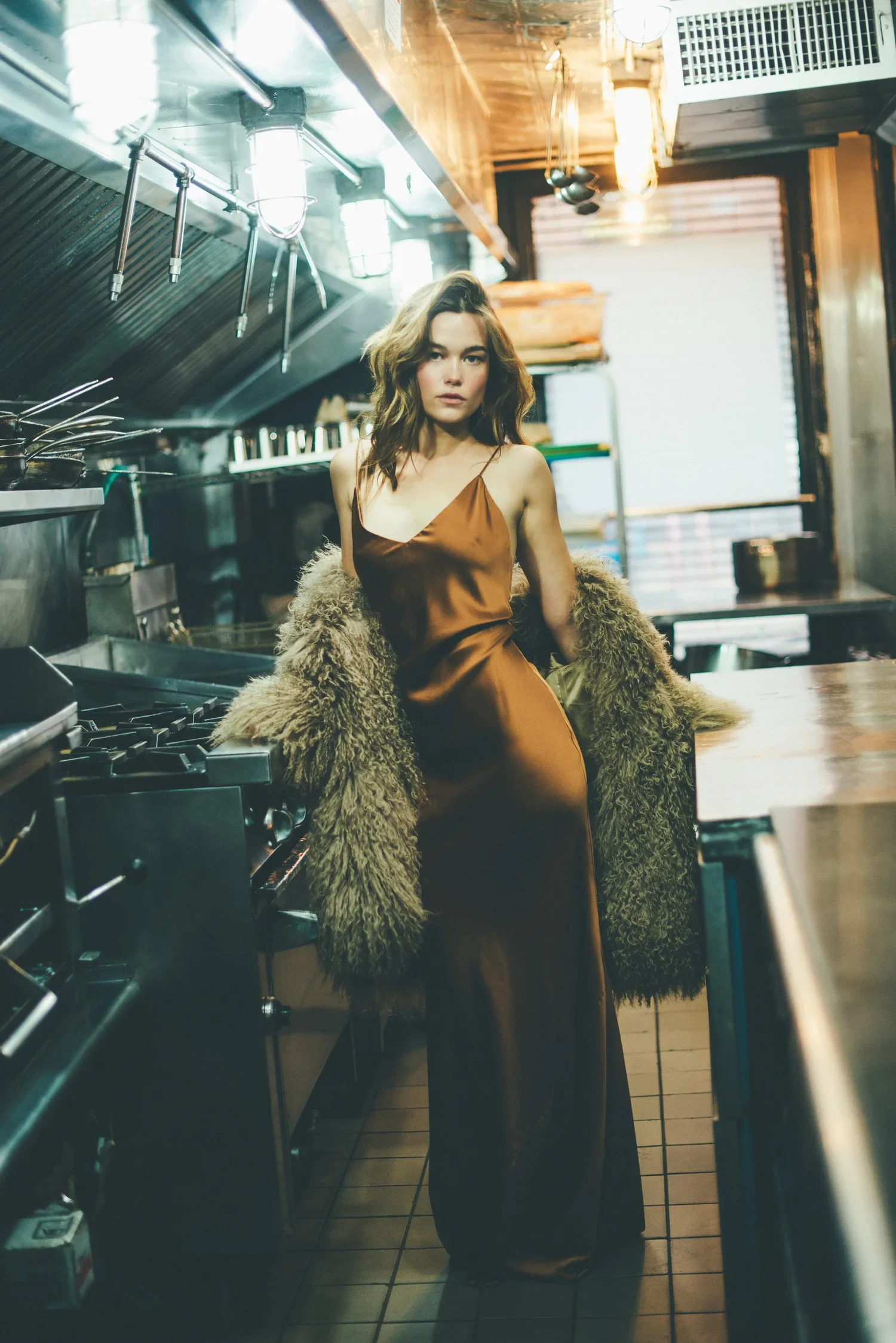 A young woman with wavy brown hair wearing a brown satin dress and a faux fur coat inside a restaurant kitchen, standing next to a stove and looking at the camera.
