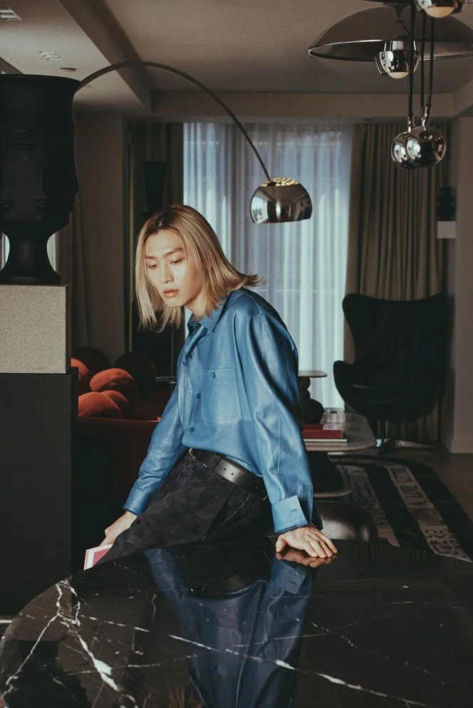 A young woman with blonde hair in a blue satin shirt and black pants leaning on a black marble table in a modern living room.