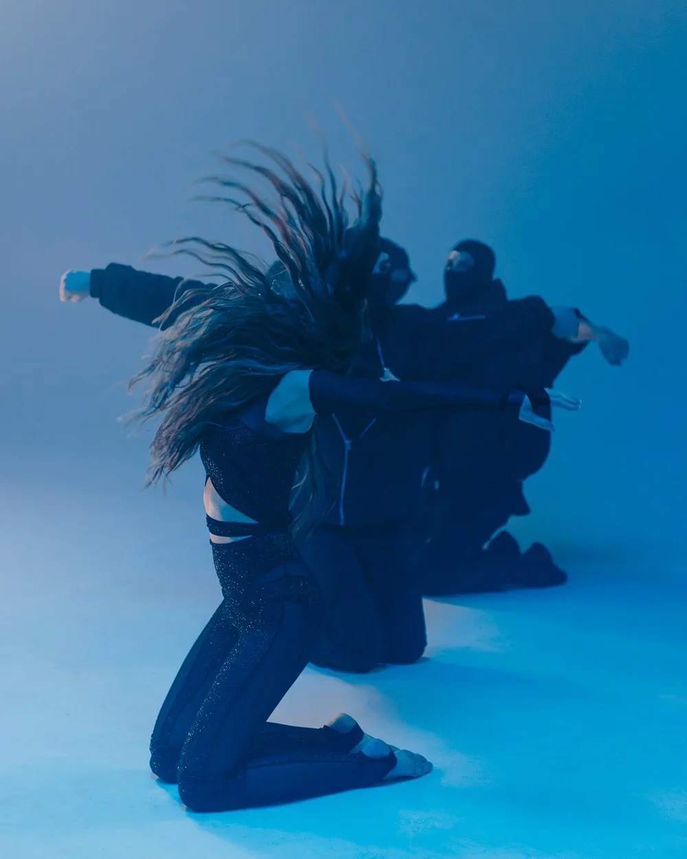 Two dancers with long hair, dressed in dark clothing, performing in a dimly lit space with blue lighting, kneeling on the floor with arms extended.