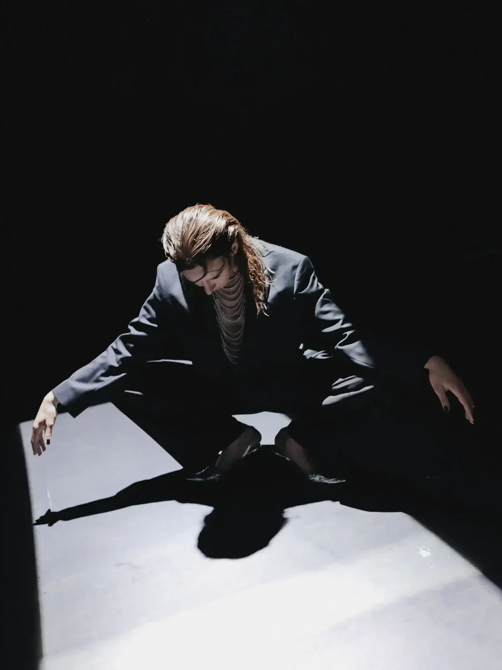Person in a black suit crouched in a dark room with lighting highlighting their figure and casting a shadow.