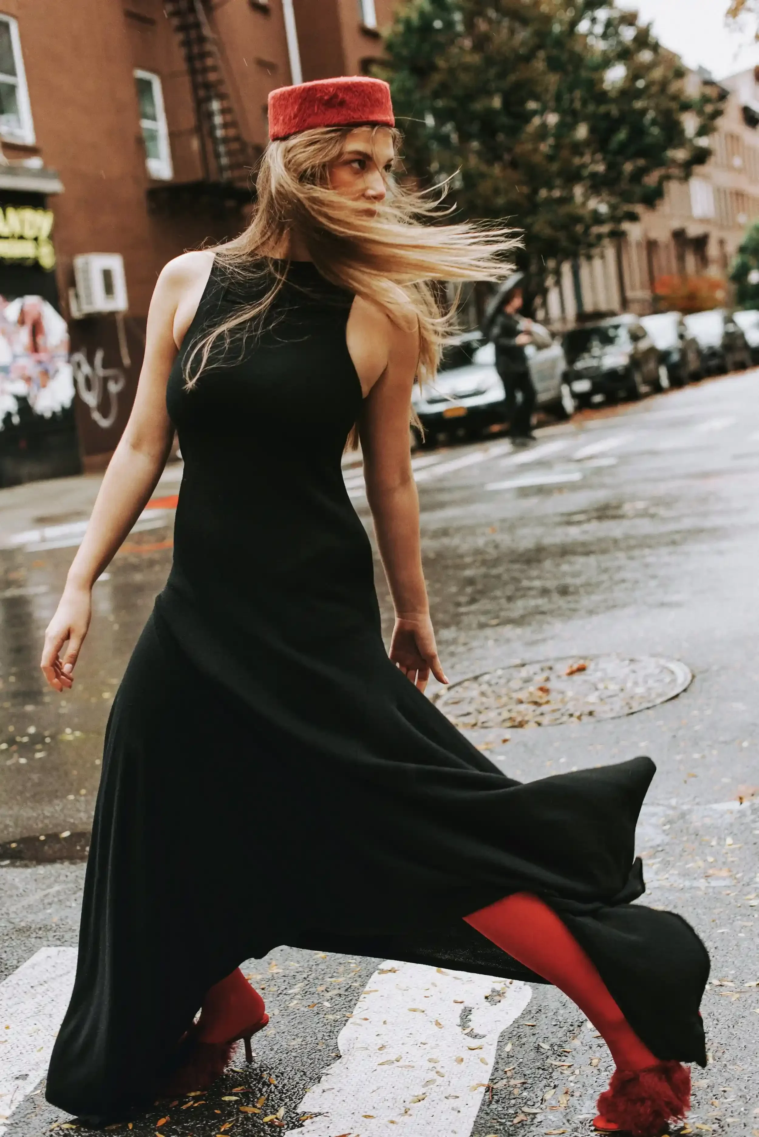 A woman in a black dress and red furry hat and red furry shoes stands on a city street with wet pavement and fallen leaves, with buildings, parked cars, and a man in the background.