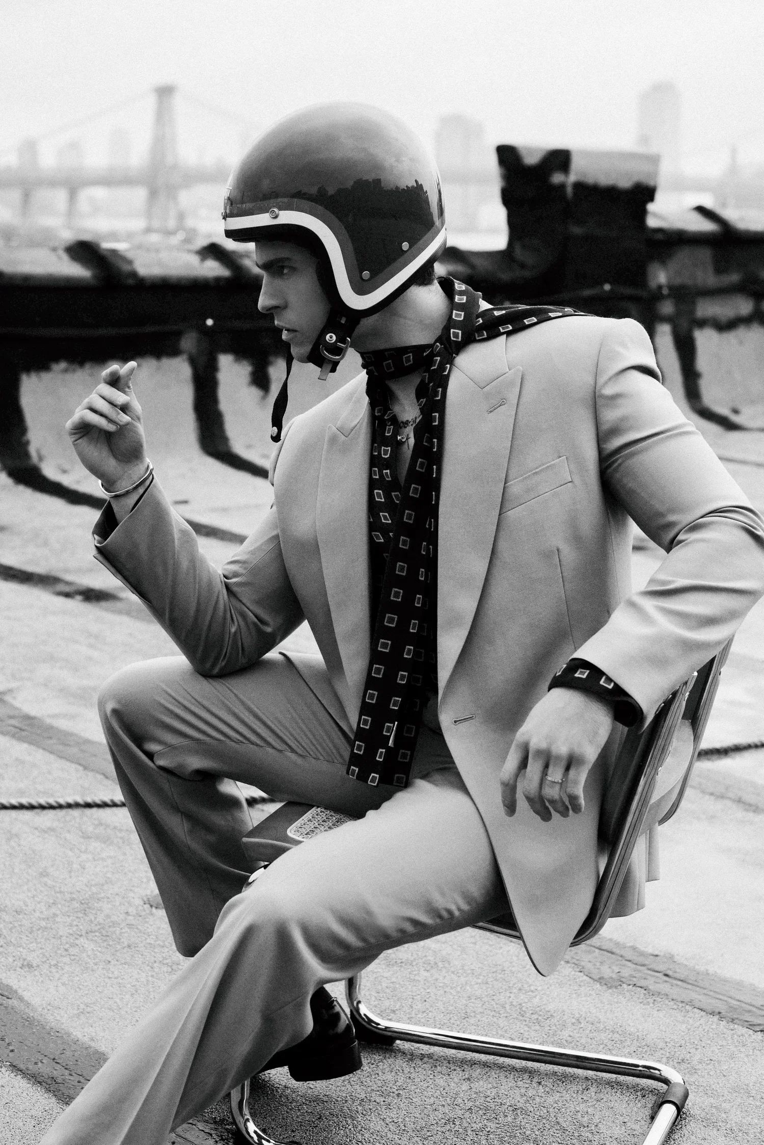 A young man sitting on a chair on an outdoor rooftop with a city skyline and bridge in the background, wearing a helmet and a blazer, looking focused.