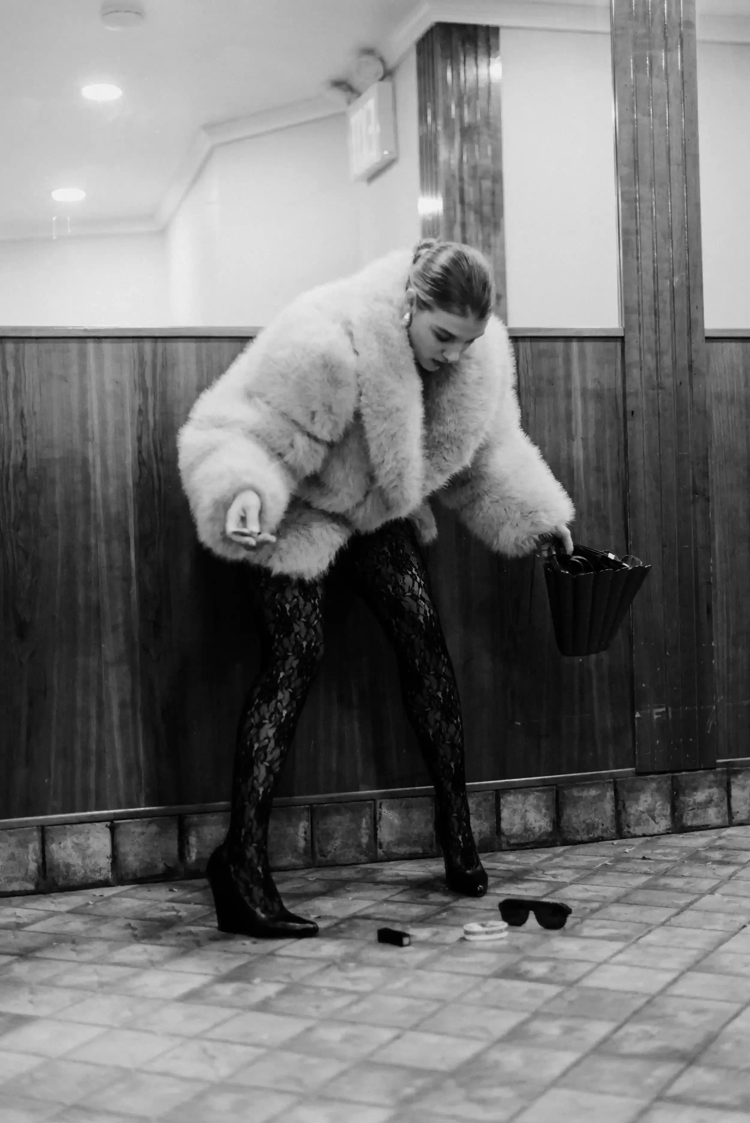 A woman with styled hair, wearing a fluffy coat and patterned tights, is standing in front of a wooden wall in a room with a tiled floor. She is looking down and holding a handbag. Scatter on the floor in front of her are a pair of sunglasses, a lips