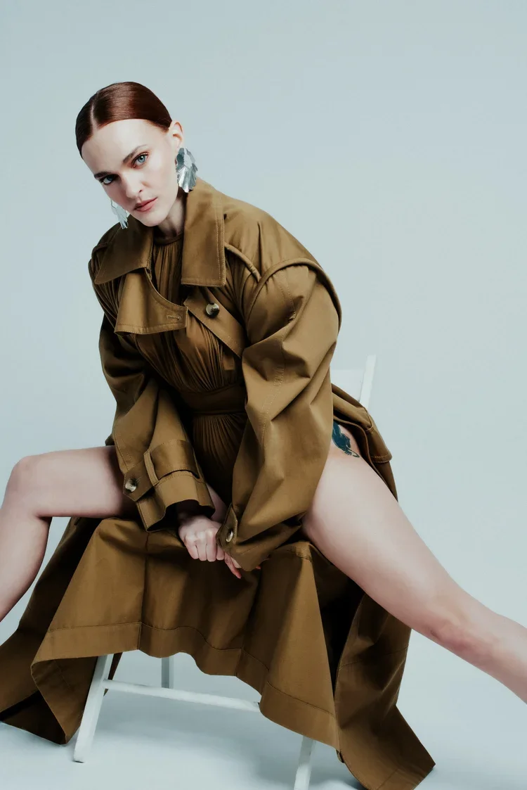 A woman with short reddish hair and large earrings, wearing a brown trench coat, poses sitting on a white chair with her legs spread apart. The coat is open, revealing a tattoo on her upper thigh. The background is plain and light-colored.