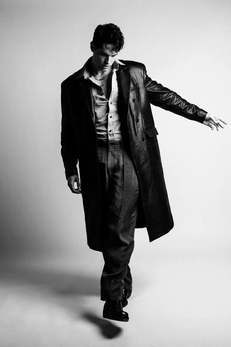 A young man in a black leather trench coat, dress shirt, and pants, walking or posing in a black-and-white studio shot with a plain background.