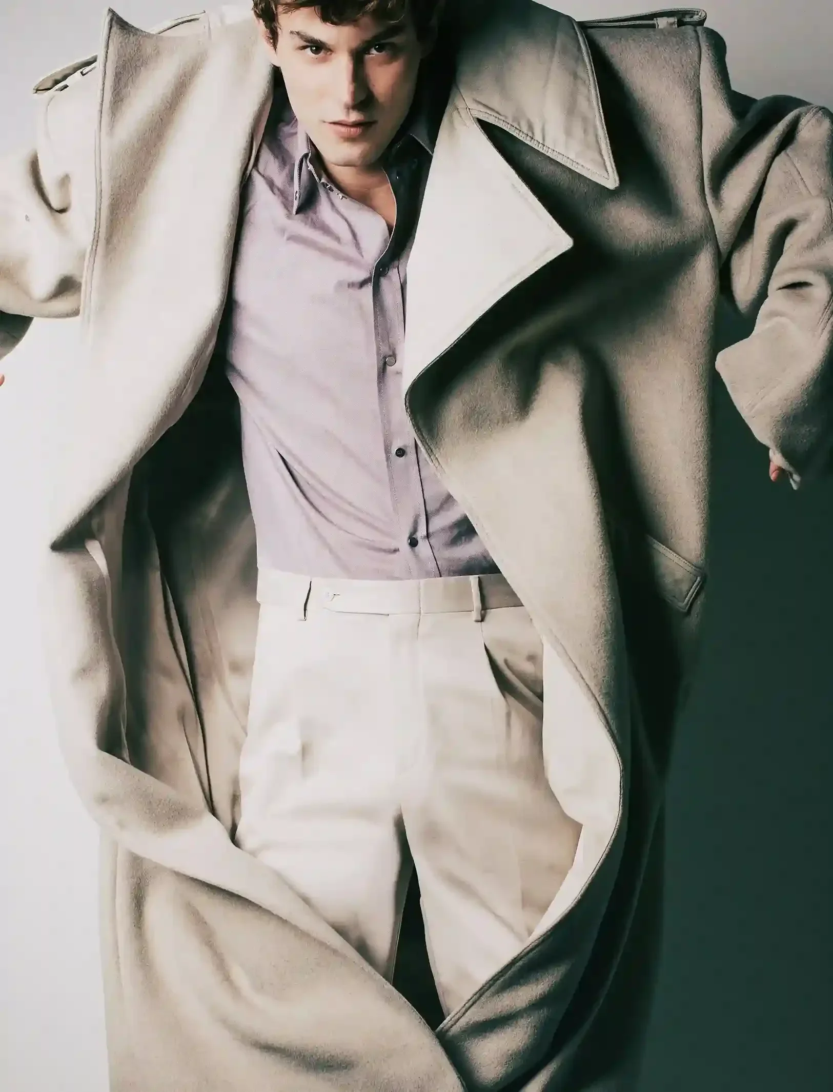 A young man with dark hair and light skin, wearing a light-colored shirt and beige pants, is leaning over the open lid of a black and white photographic print of himself that is placed on a flat surface. The man appears to be gazing intently at the c
