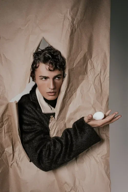 Person with short curly hair looking through a torn piece of brown paper while holding a white egg in their hand.