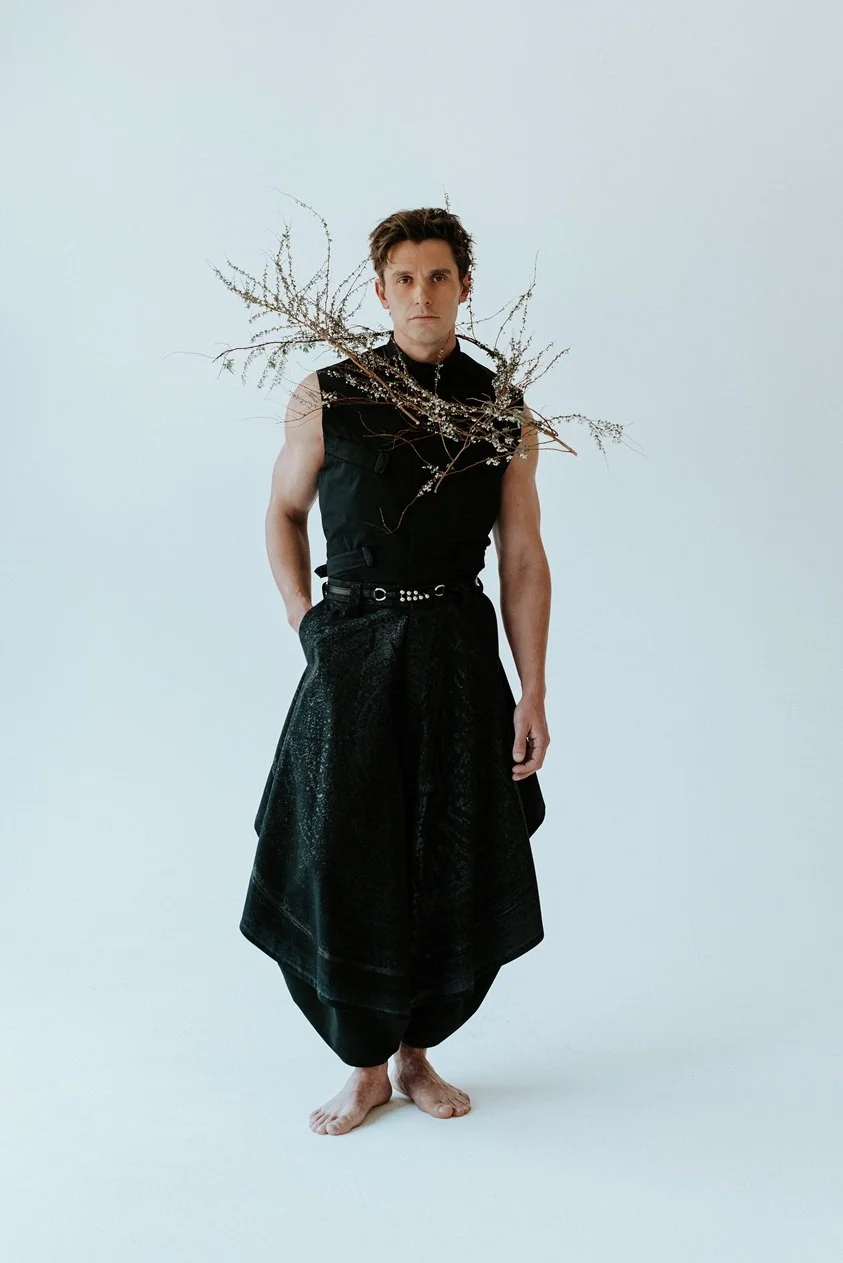 A man standing barefoot in a studio, wearing black clothing with dried branches draped across his shoulders, looking directly at the camera against a plain light background.