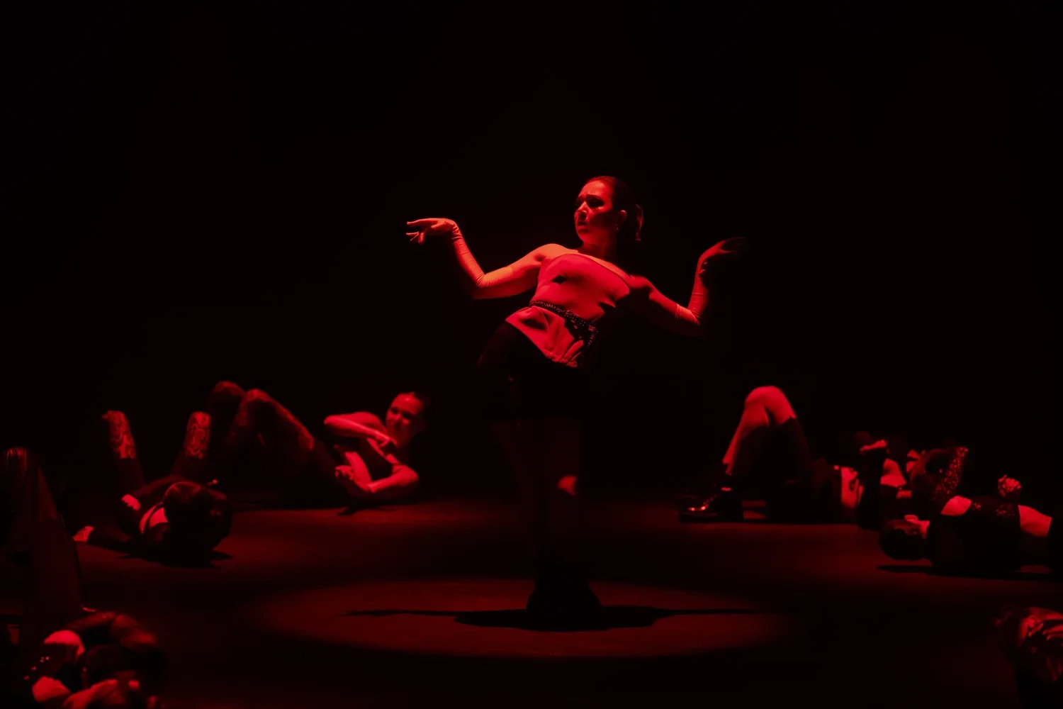 A female dancer performing on stage illuminated by red lighting, surrounded by other dancers lying on the floor in various poses.