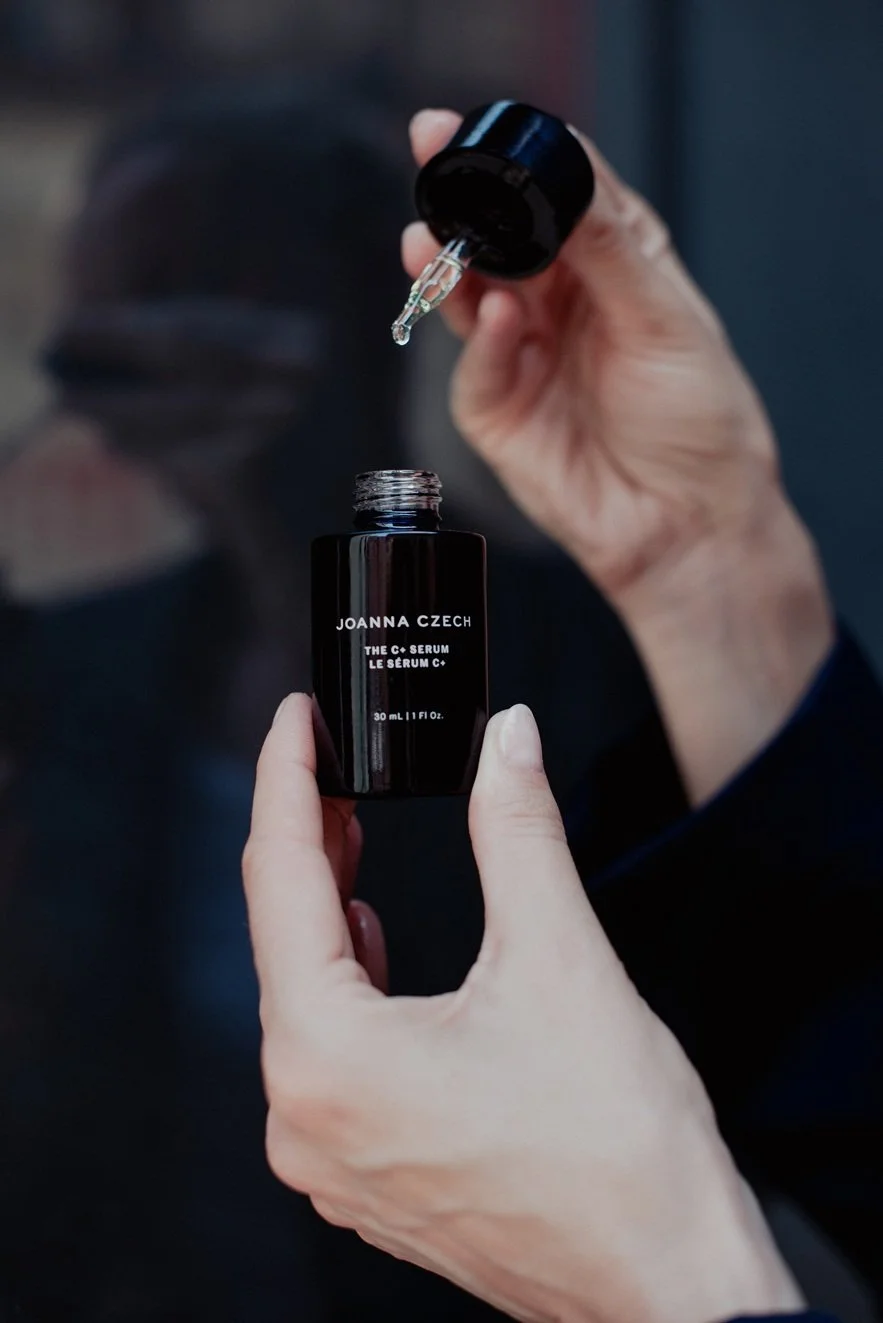 Person holding a black dropper bottle labeled 'Joanna Czech, The C+ Serum,' with a drop of serum about to be dispensed.