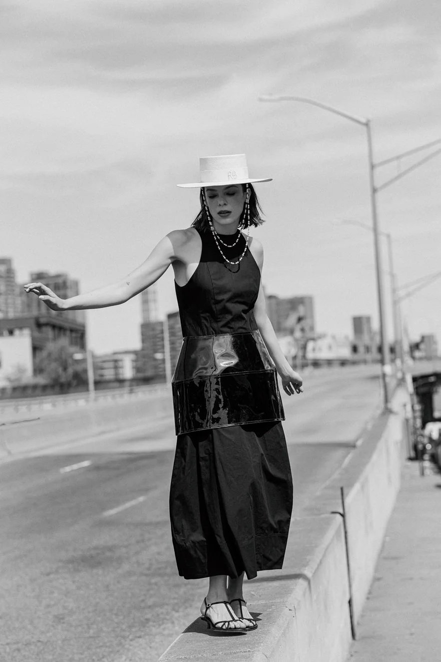 A woman in a stylish black dress and a wide-brim hat walking on a city sidewalk with tall buildings in the background, captured in black and white.