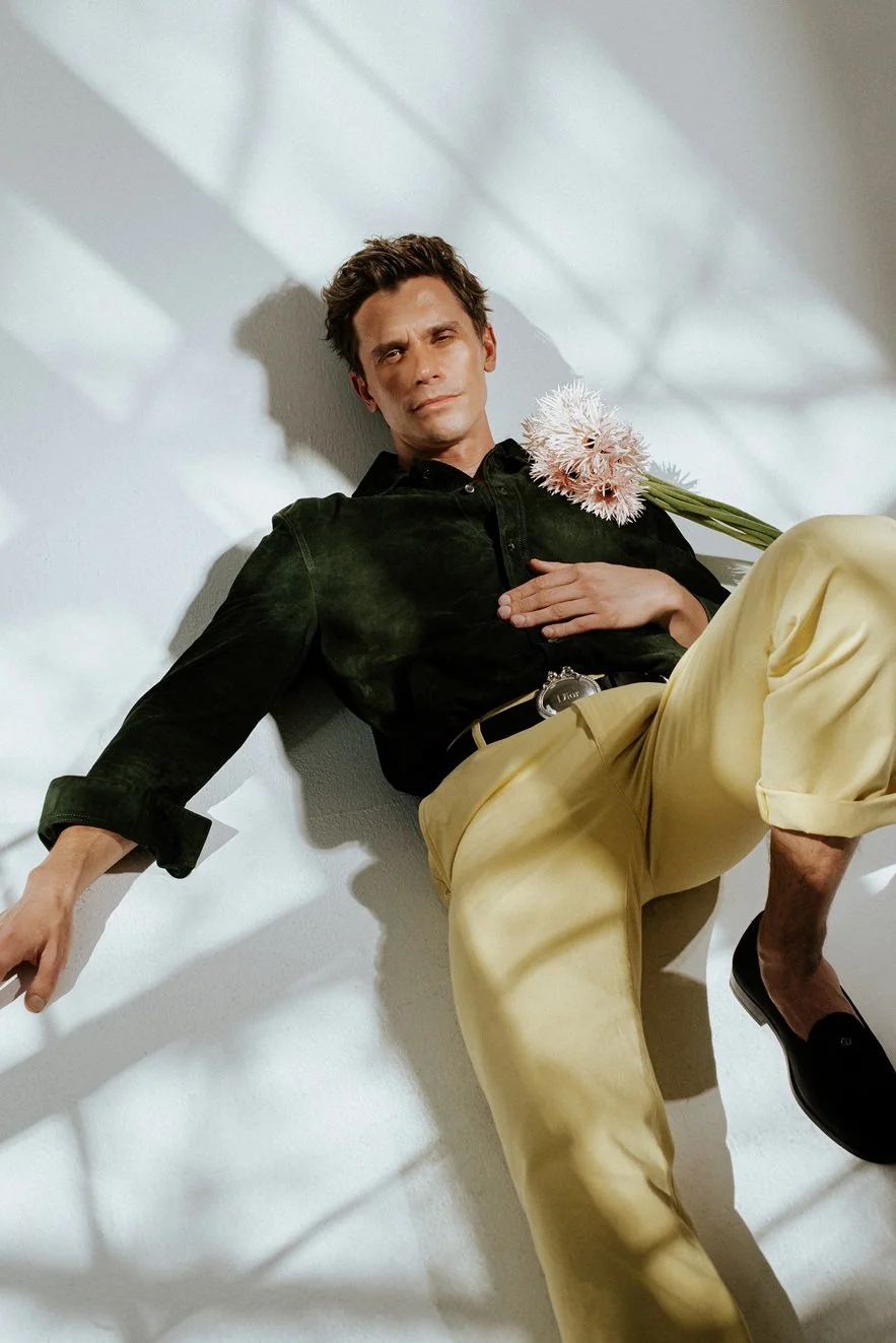A man with dark hair reclining on the floor with a pink flower on his chest, wearing a black shirt and beige pants, with shadows from window blinds on the wall behind him.