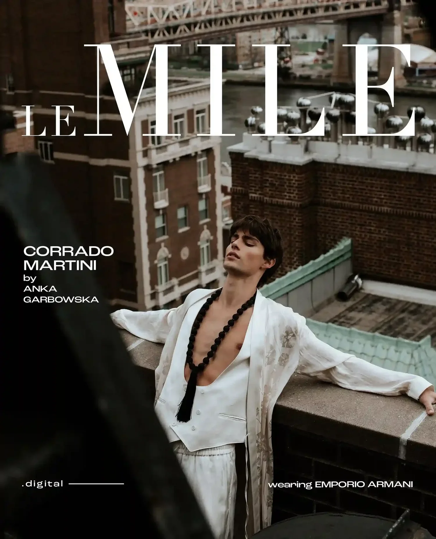A young man with dark hair and a fair complexion is standing on a rooftop with his arms outstretched and eyes closed. He is wearing a loose, white, patterned robe with a deep V-neck, black beaded necklace, and matching earrings. The background shows 