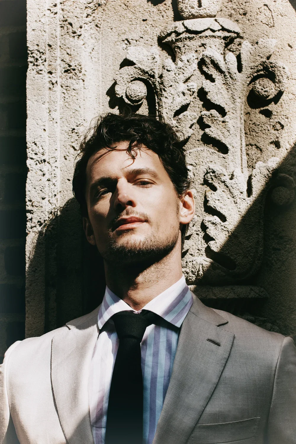 A man wearing a light-colored suit, striped shirt, and black tie standing against an ornate stone wall with carved designs, with sunlight casting shadows.