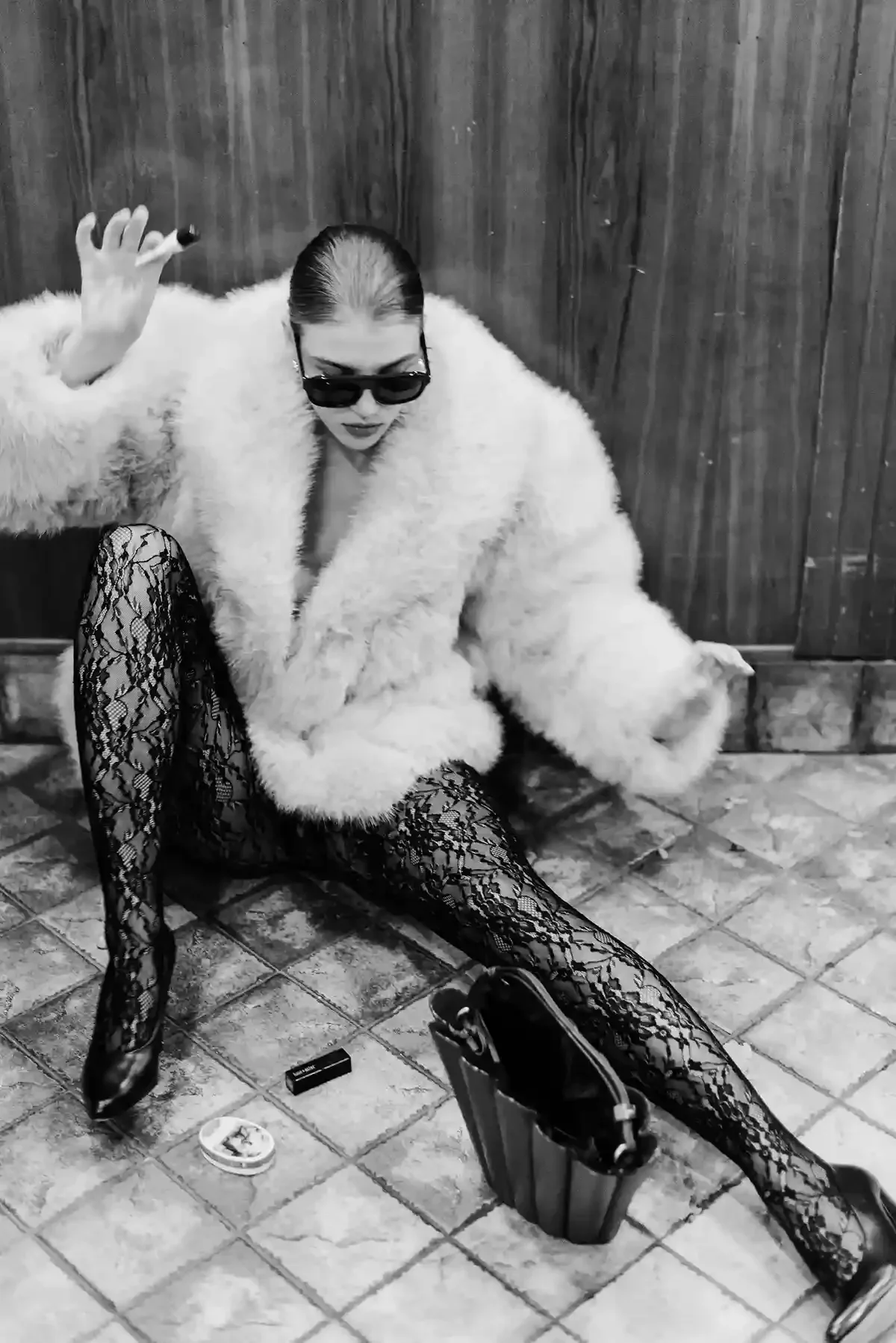 A woman wearing sunglasses, a fuzzy coat, and lace tights, sitting on a tiled floor against a wooden wall, with her legs spread apart, holding a cigarette, a handbag on the floor, and makeup items nearby.