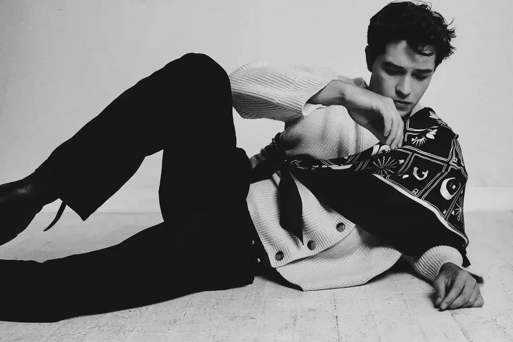 A young man lying on the floor in a relaxed pose, holding a bandana over his shoulder, dressed in a long-sleeve shirt and black pants.