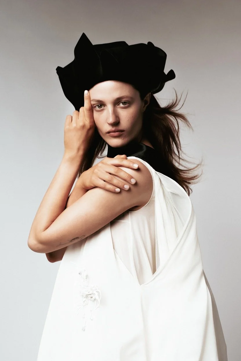 A young woman with fair skin and brown hair wearing a large black hat and a white sleeveless dress standing against a plain gray background. She is touching her forehead with her right hand and resting her left hand on her shoulder.