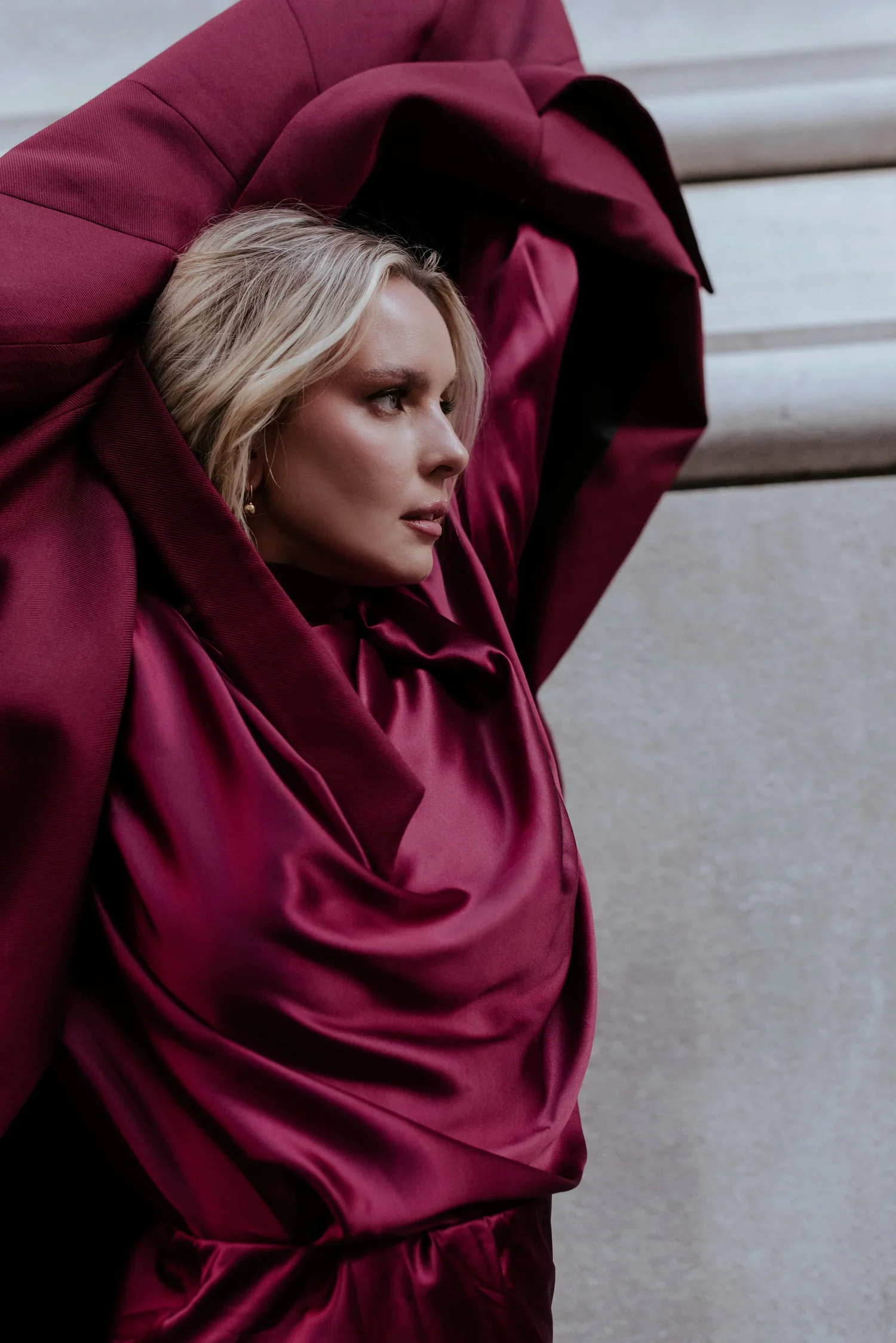 A woman with blonde hair in a low bun, wearing earrings and a burgundy satin coat, is lying on a light gray concrete surface with wooden steps in the background.