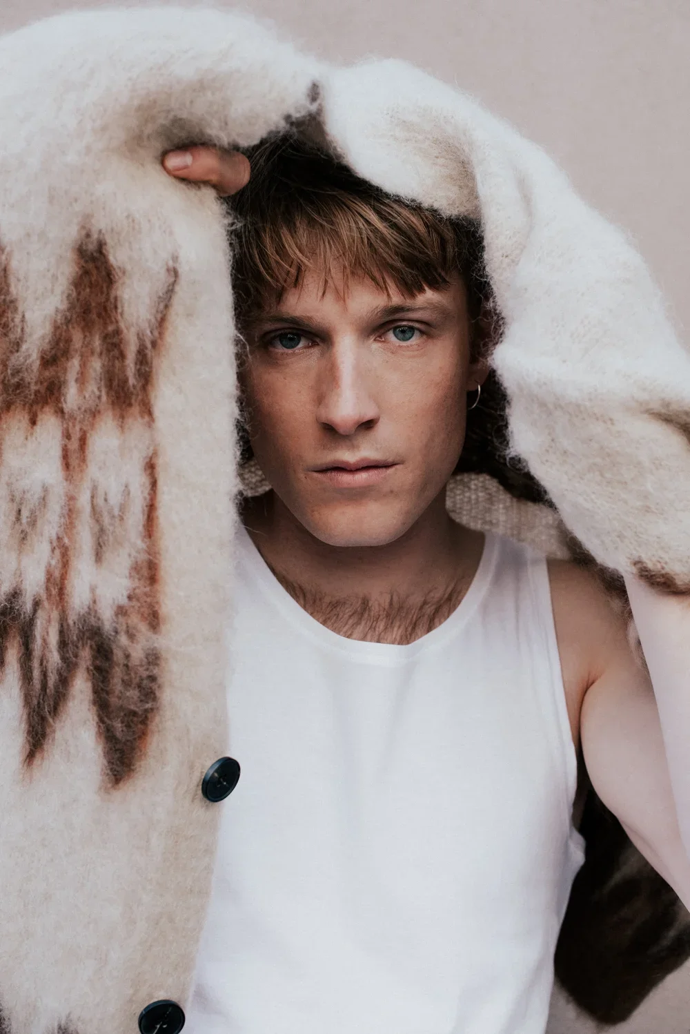 A young man with blue eyes and brown hair, wearing a white sleeveless shirt, is holding a large, fluffy, beige and brown fur coat over his head.