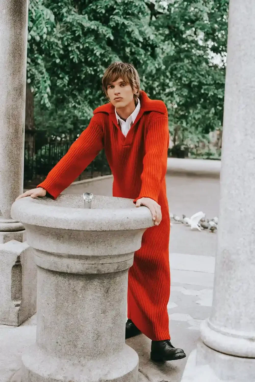 Young man in red sweater leaning on stone drinking fountain in outdoor park setting with green trees and white birds.