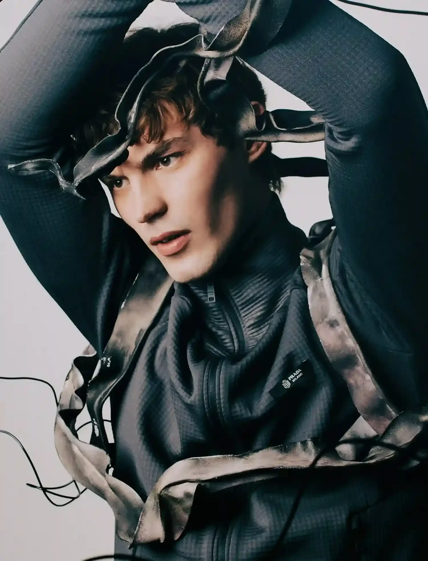 A young man with dark curly hair and fair skin wearing a black Prada jacket, looking thoughtfully while holding a pair of black sunglasses above his head, seated against a plain light background.