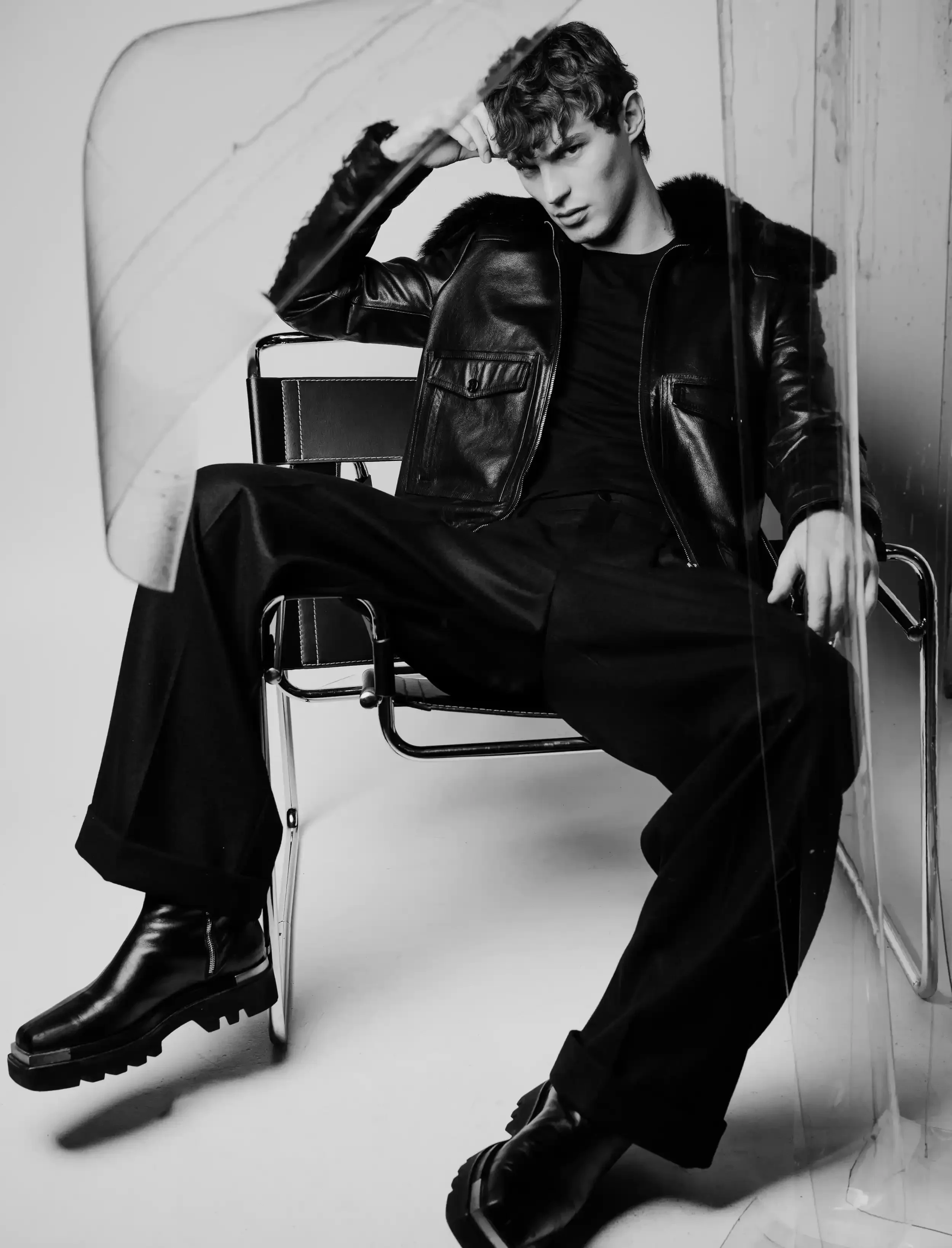 Black and white photo of a young man with curly hair wearing a leather jacket, sitting on a chair with one arm resting on his head, leaning against a wall, with a serious expression.
