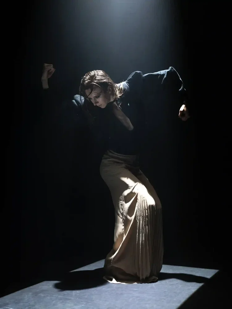 A woman with dark, wet hair and dark clothing, striking a dramatic pose in a spotlight, against a dark background.