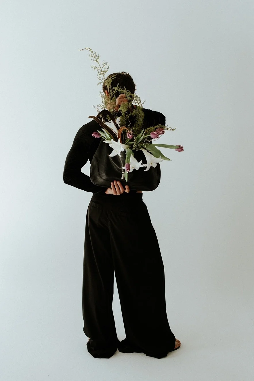 Person holding a black handbag with a large floral arrangement of lilies and tulips, obscuring their face, standing against a plain light background.