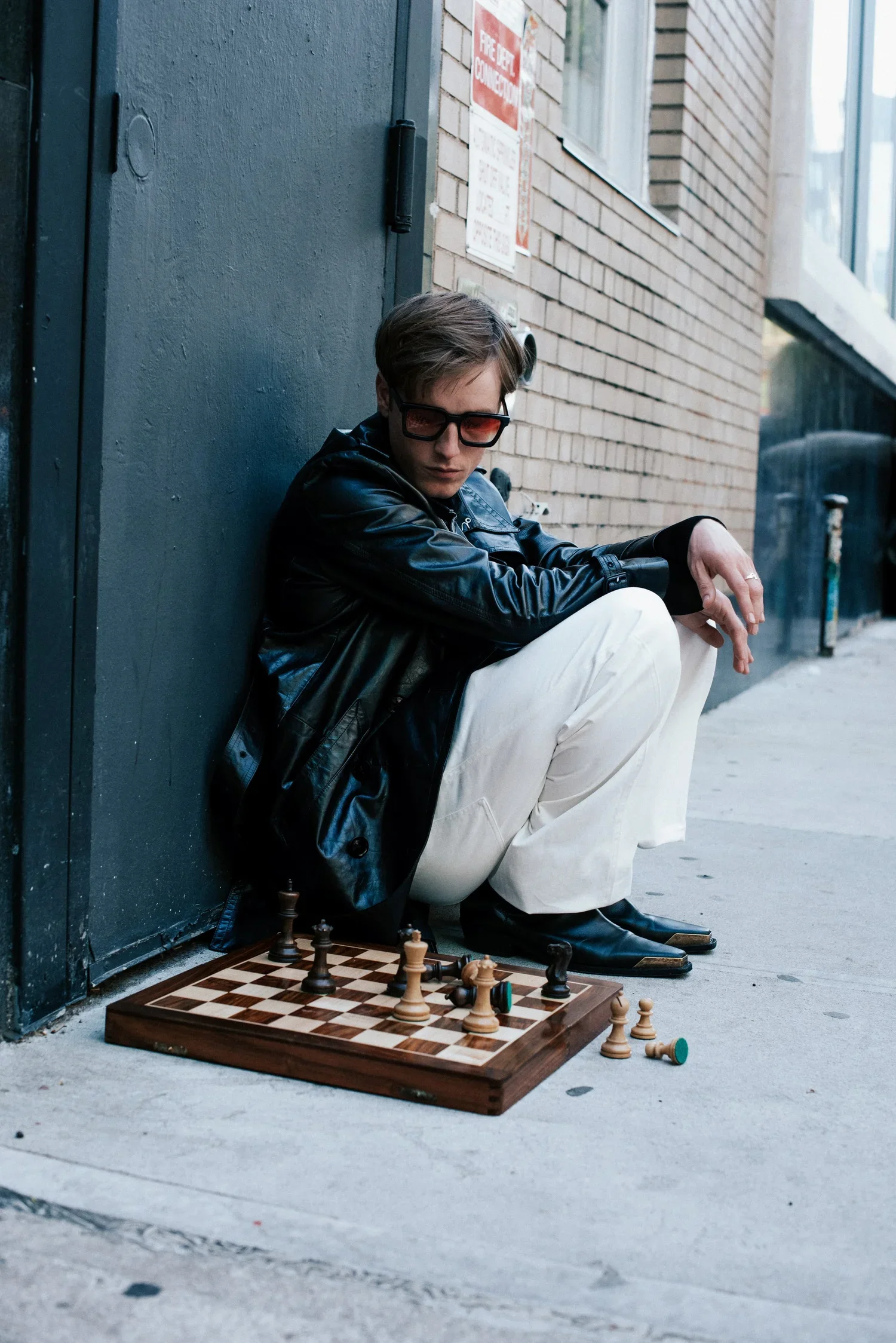 A young person with short hair, wearing sunglasses, a leather jacket, and white pants, sitting on the sidewalk against a dark blue door and brick building, playing chess on a wooden board with chess pieces nearby.
