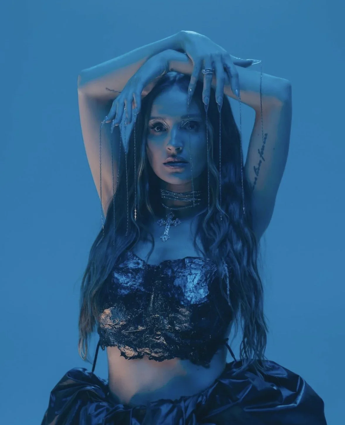 A woman with long wavy hair, wearing dark clothing, jewelry, and tattoos, poses with her arms raised above her head against a blue background.