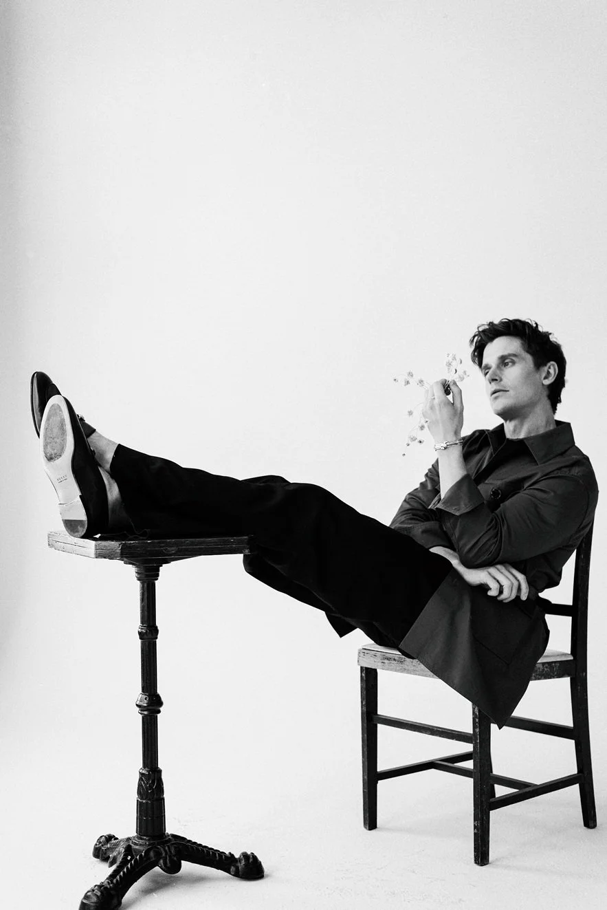 A black and white photograph of a man with dark hair, sitting on a wooden chair with one leg propped on a small, ornate table. He is dressed in a dark shirt and pants, holding a cigarette, and appears to be in a contemplative pose against a plain whi