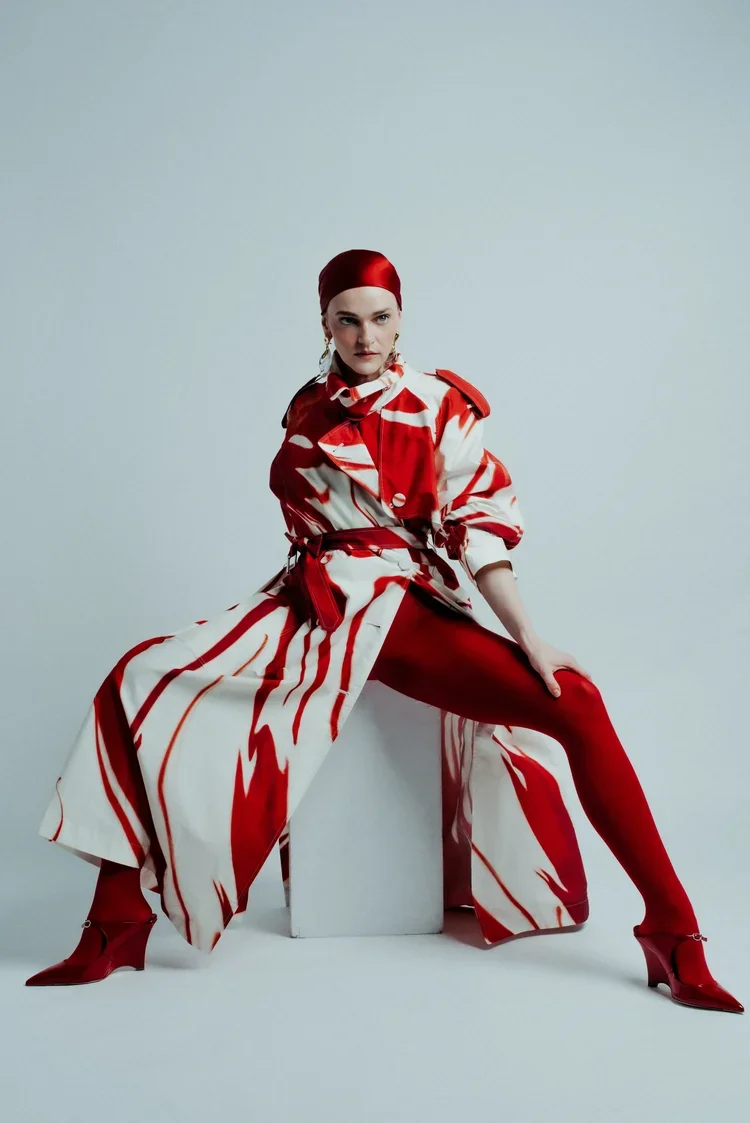 A woman wearing a red and white patterned trench coat, red tights, and red high heels, sitting on a white block against a plain gray background.