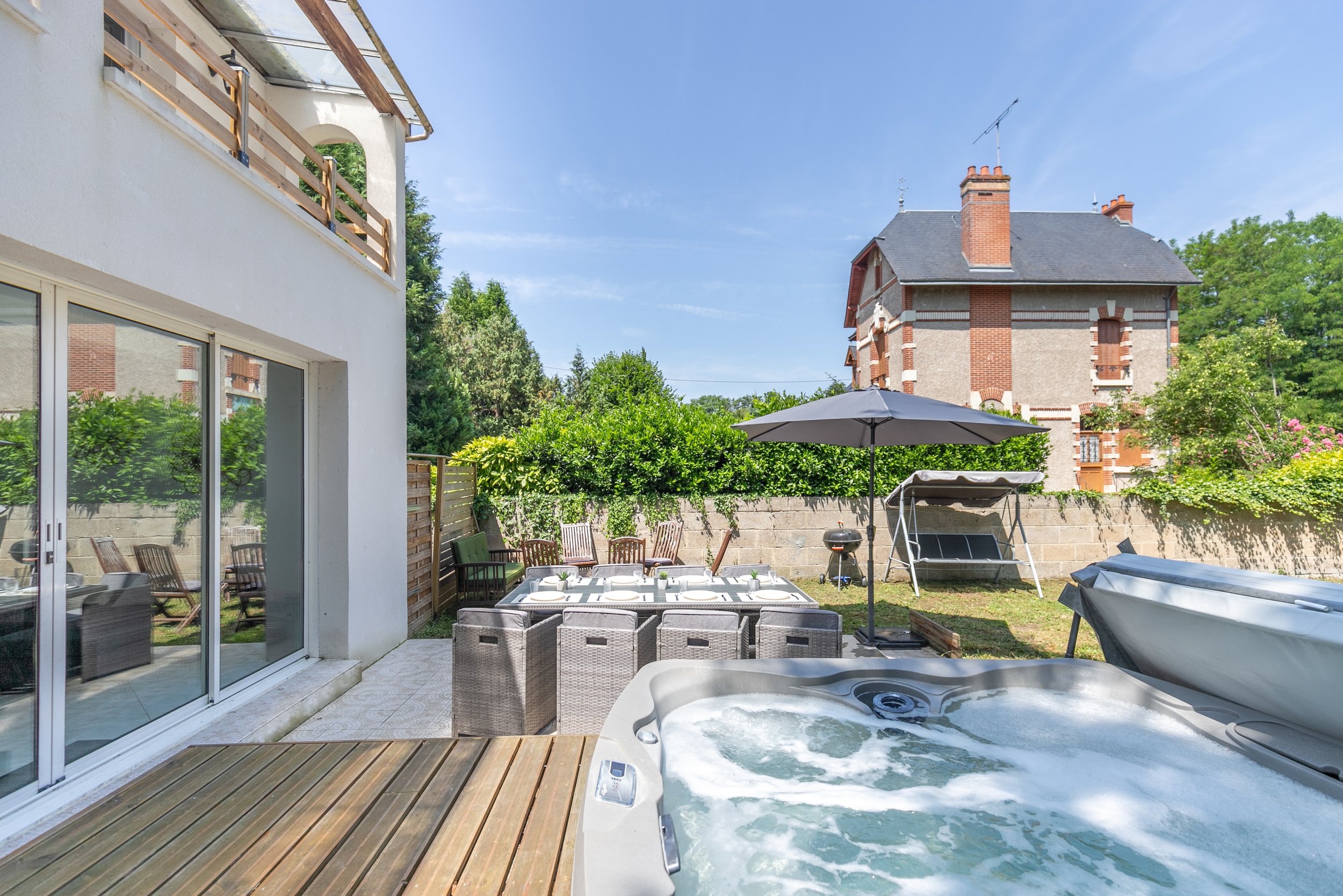 Jardin de maison avec jacuzzi, table pour repas, parasol, chaises, barbecue, balancelle, et maison en arrière-plan