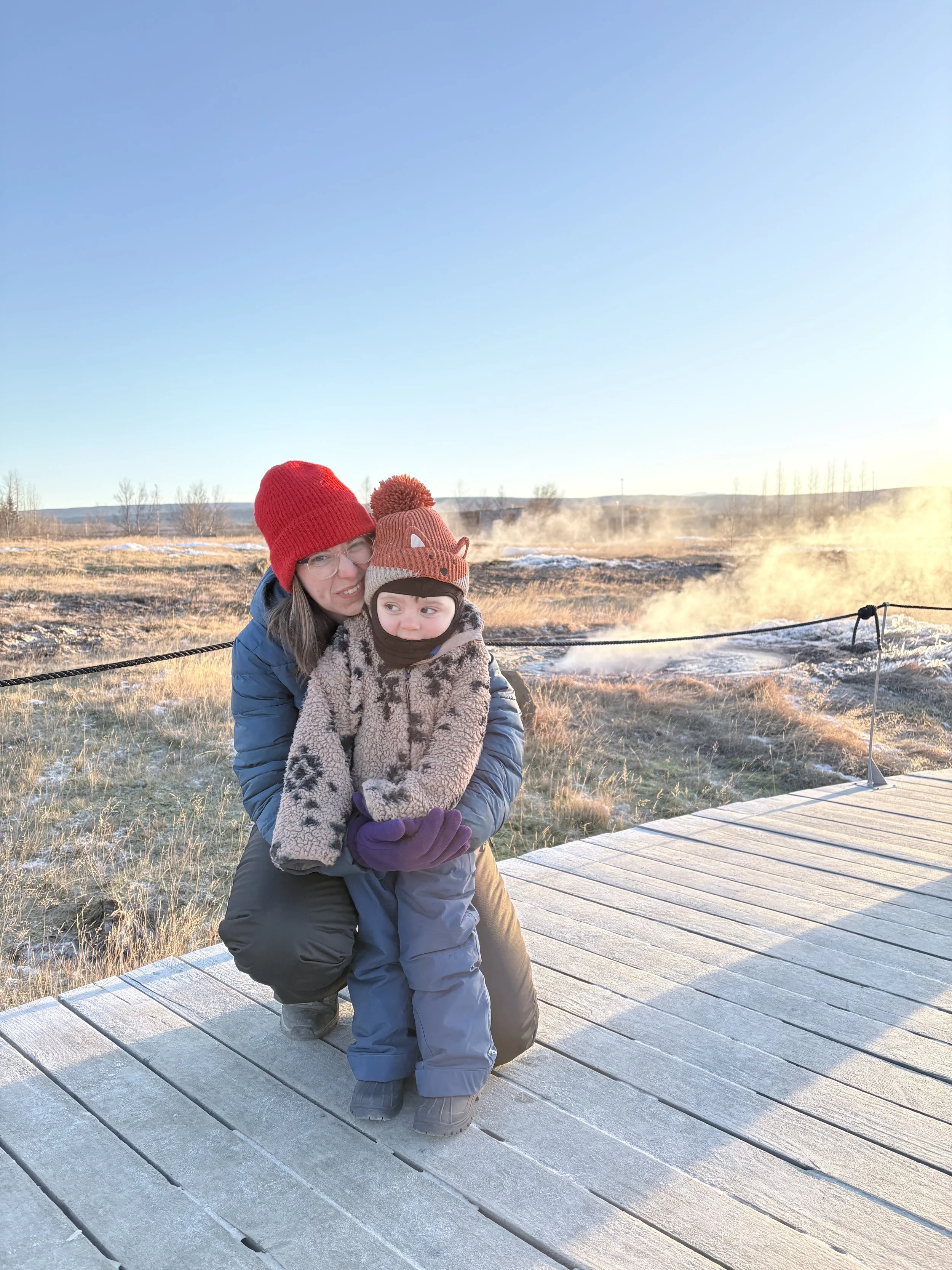 A woman in a red beanie and glasses hugging a young child in a warm hat and coat on a wooden boardwalk in a cold, steaming landscape.