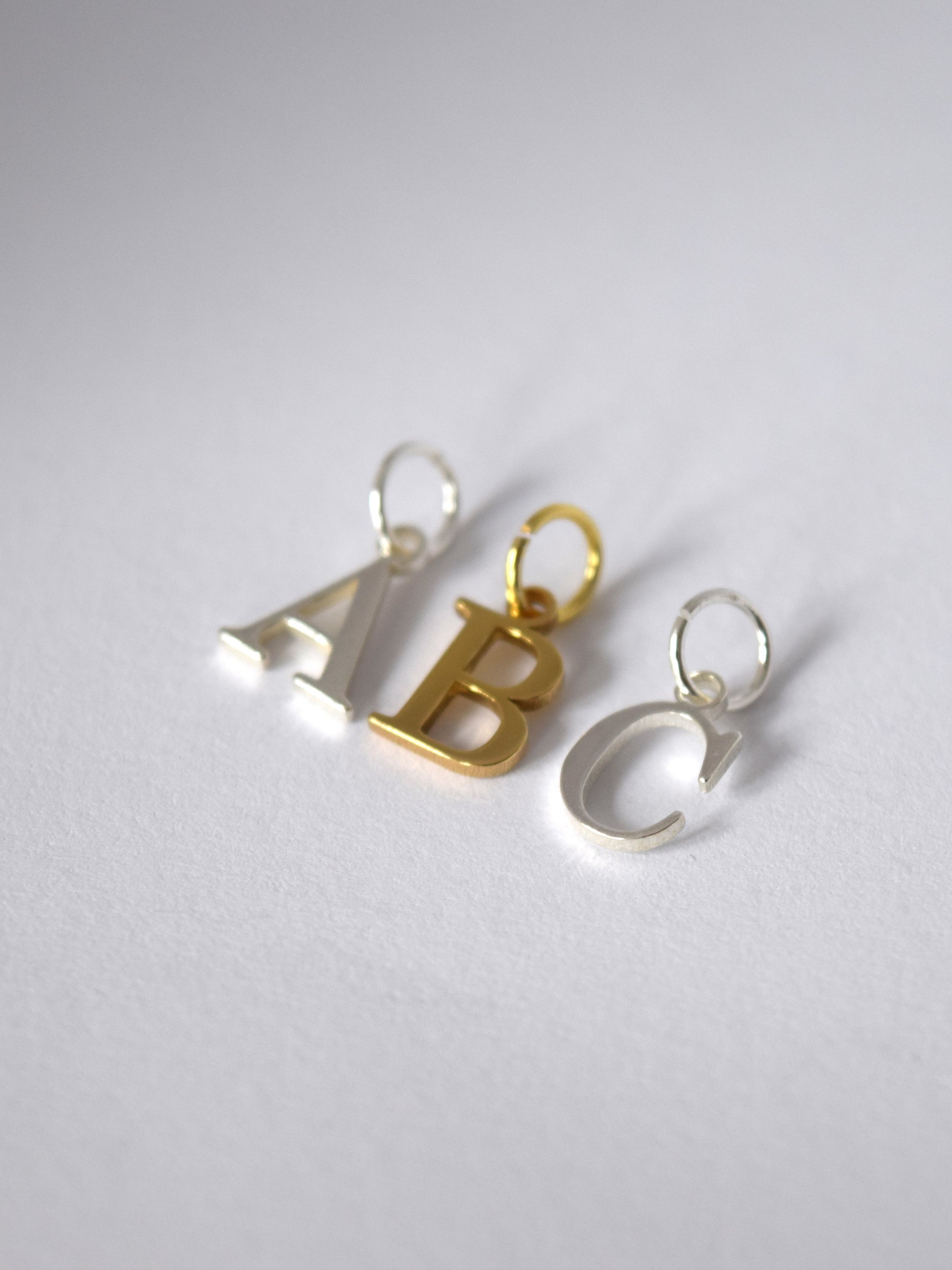 Close-up photo of three letter-shaped pendants spelling 'A', 'B', and 'C'. The 'A' and 'C' pendants are silver, and the 'B' pendant is gold. Each pendant has a small circular loop at the top for attaching to a chain or bracelet, and they are arranged on a white surface with soft shadows.