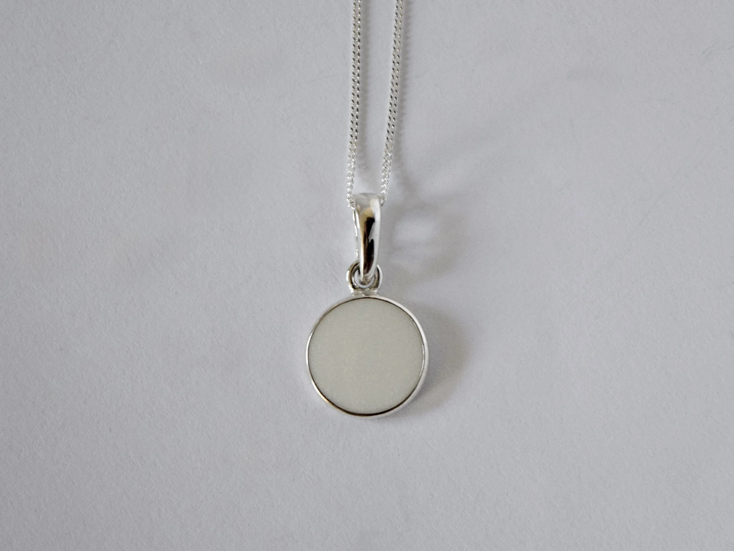 A silver necklace with a round white pendant hanging against a white background.