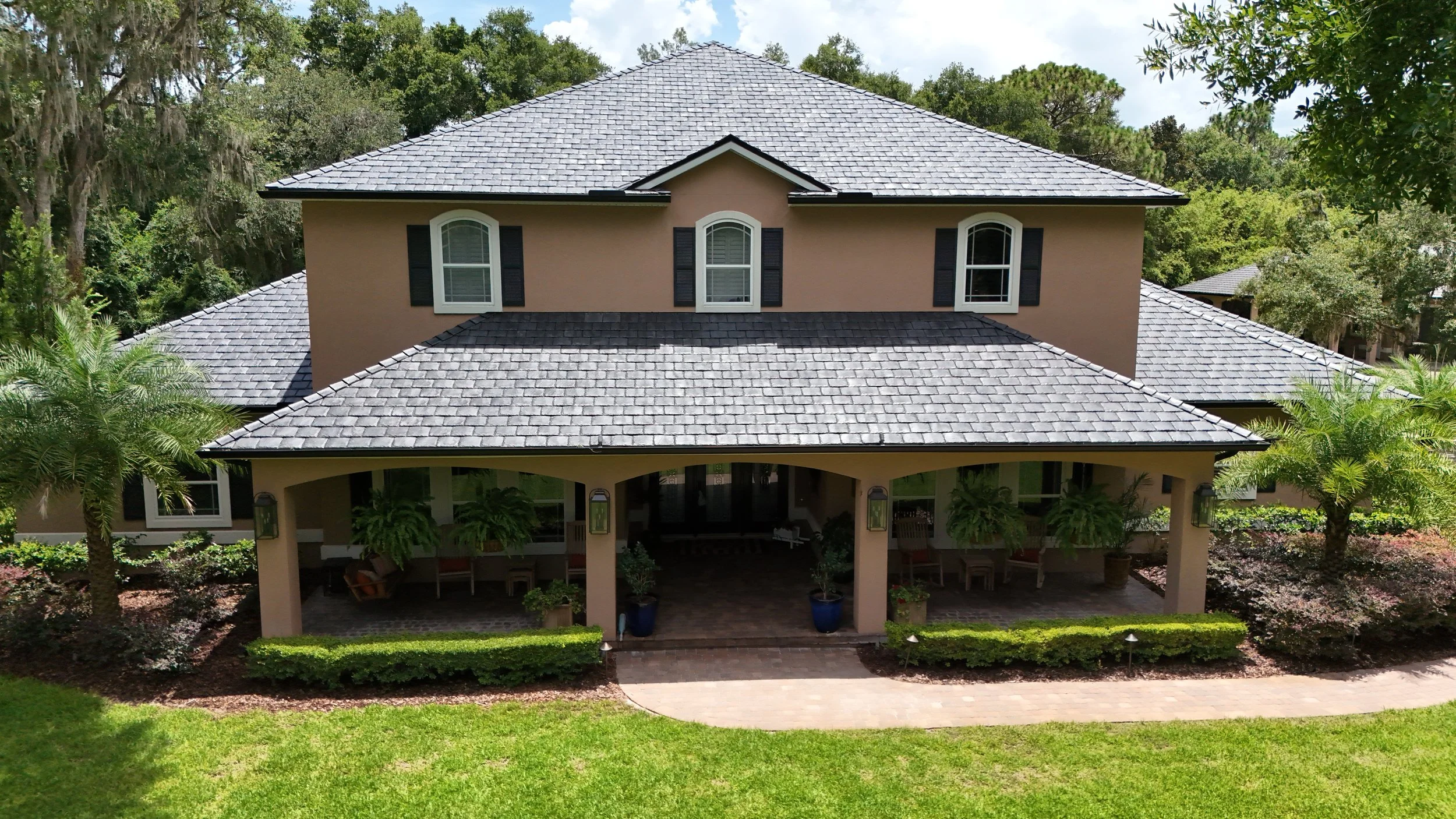 A two-story house with a gray shingled roof, beige exterior walls, and arched windows with black shutters, surrounded by green trees and bushes.