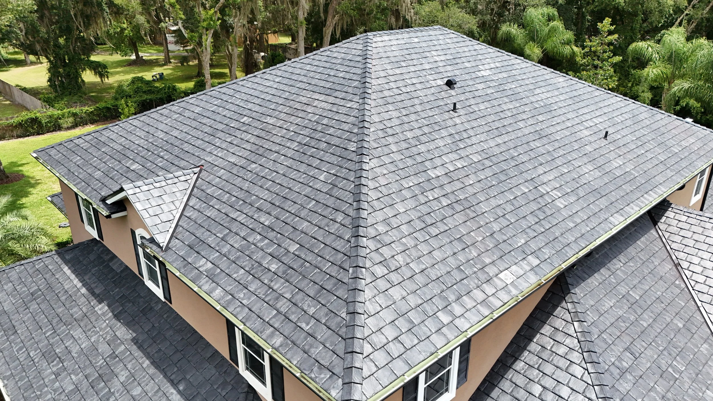 Aerial view of a house roof with gray shingles, surrounded by trees and greenery.