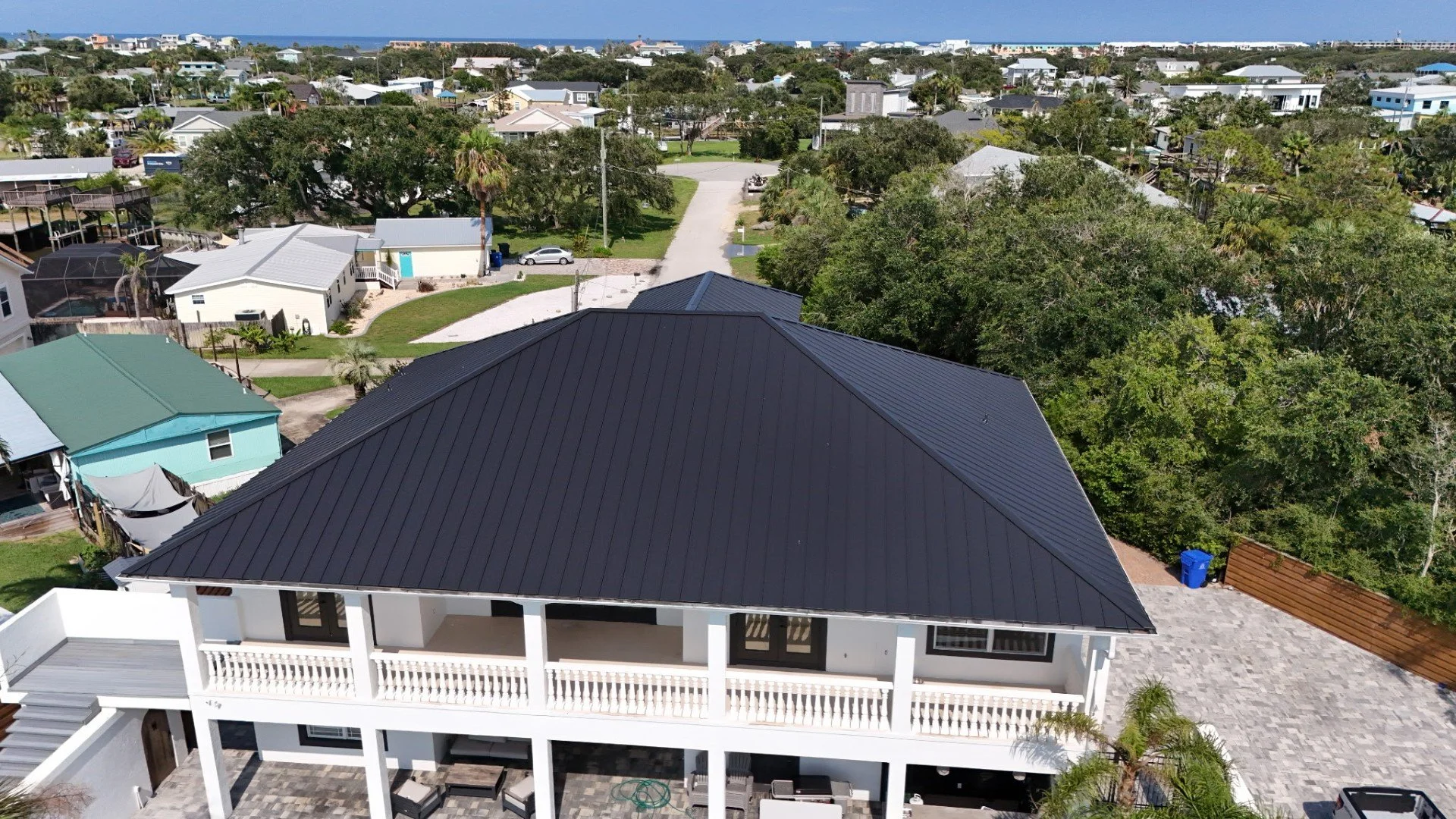 An aerial view of a neighborhood with houses, trees, and a street, with a view of the ocean in the distance.