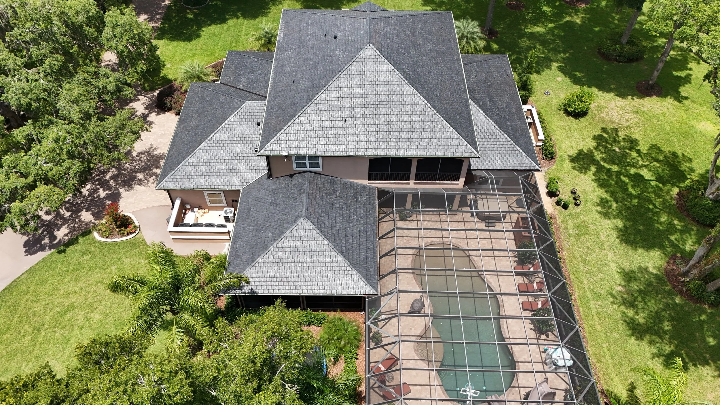 An aerial view of a backyard with a screened-in swimming pool, patio area, and a multi-story house surrounded by green trees and grass.