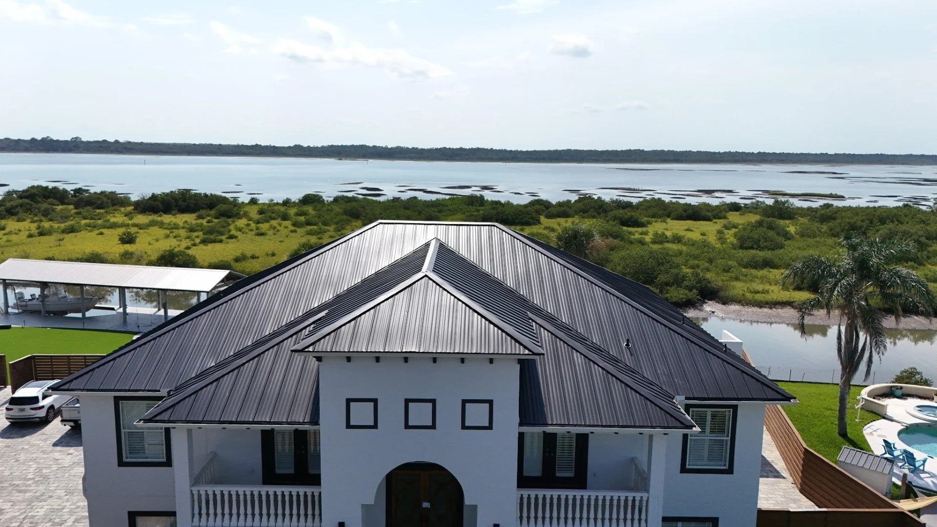 A large modern house with a gray metal roof, white exterior walls, and multiple windows, surrounded by a landscaped yard with palm trees and a swimming pool. In the background, there is a marshy landscape with water and vegetation, and beyond that, a