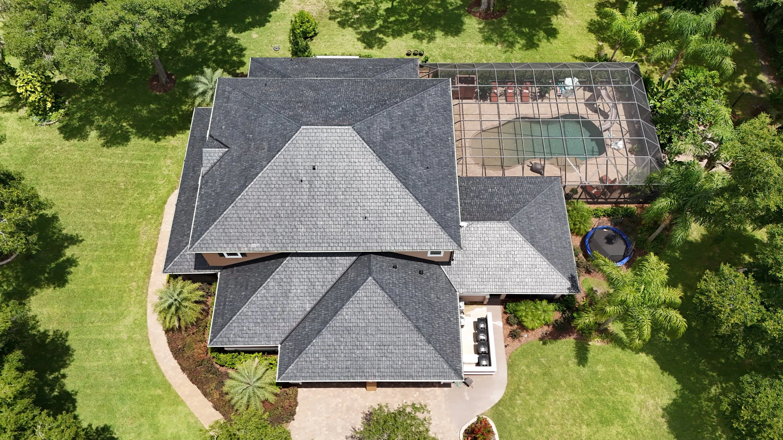 Aerial view of a house with a complex roof, swimming pool enclosed in a screened patio, trampoline, palm trees, and lush green landscape.