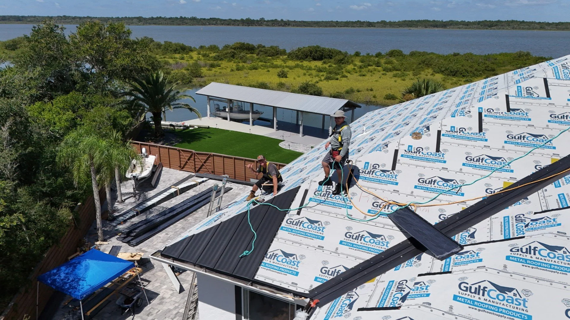 Workers installing metal roofing panels on a house, with a scenic view of a lake and lush greenery in the background.