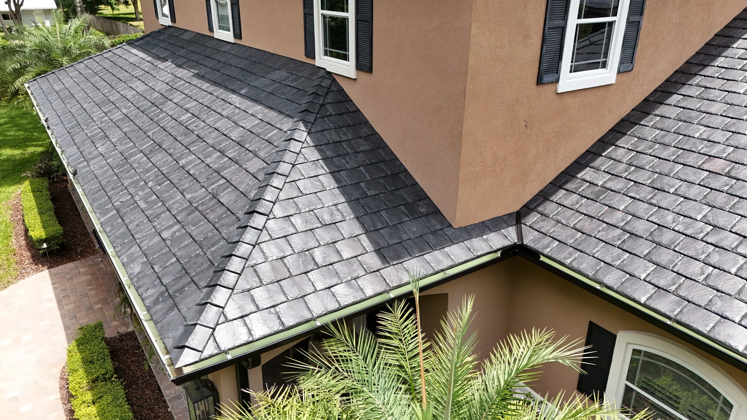 A house with a gray tiled roof and beige stucco walls, including black shutters on the windows. There is a small landscaped yard with palm trees and hedges.
