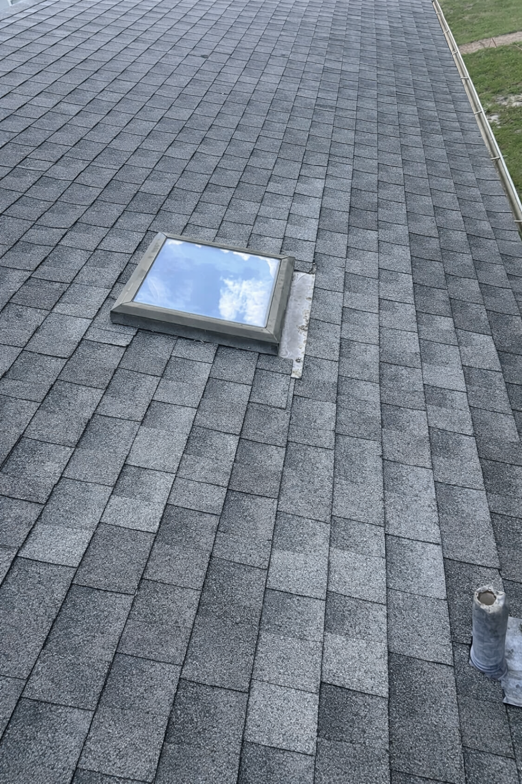 A section of a gray shingled roof with a skylight reflecting blue sky and clouds, and a nearby vent pipe.