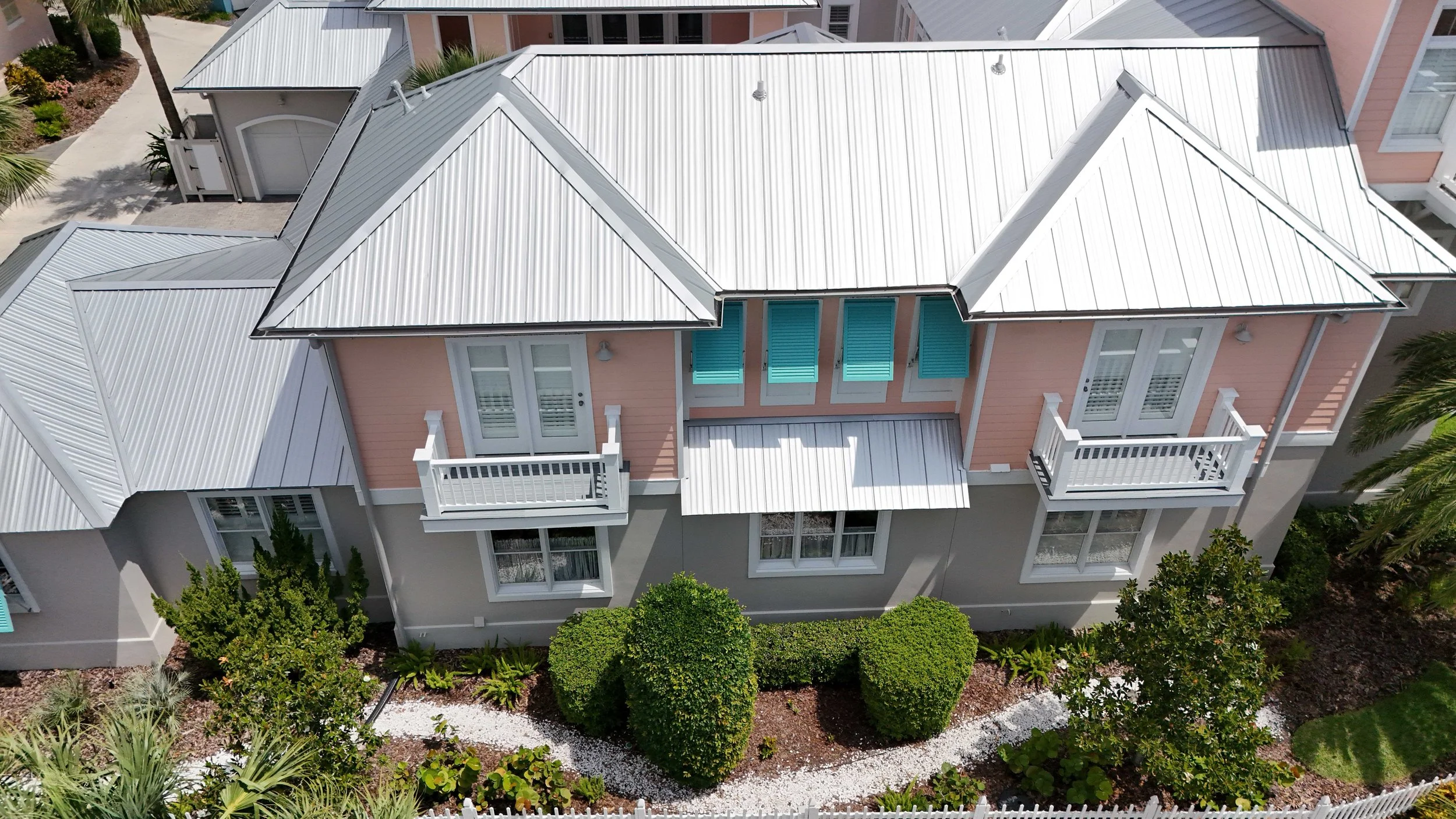 An aerial view of a multi-story house with light pink and gray exterior walls, metal roof, and small balconies with white railings. The house is surrounded by a landscaped garden with bushes, trees, and a white gravel walkway.